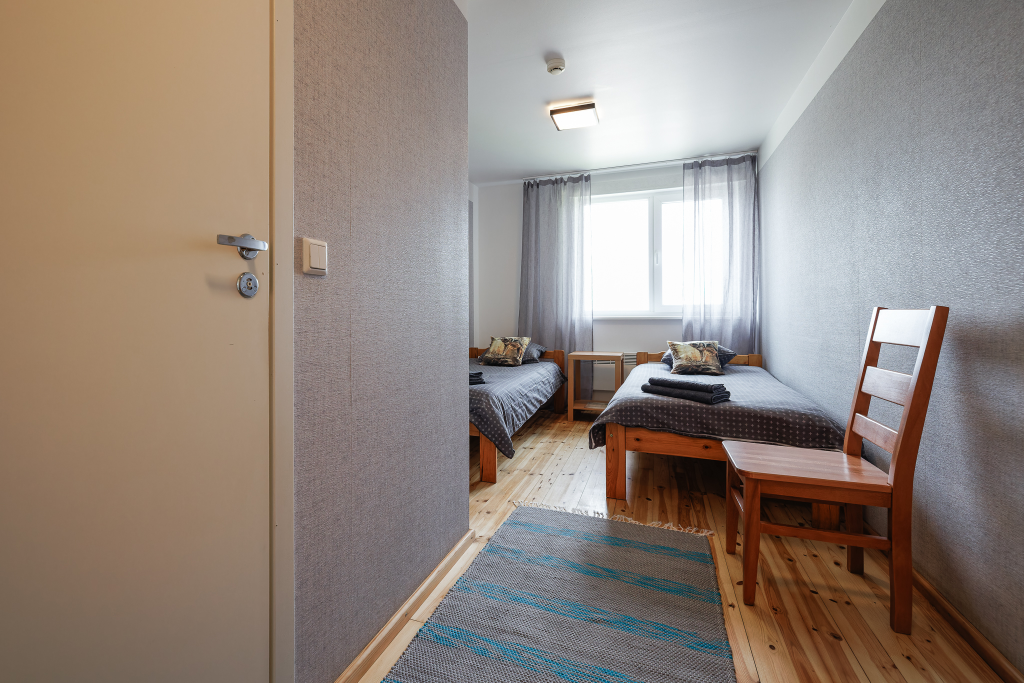 Vaade HUNT toa sissepääsust Suurlahe Guesthouse majutuses kahe eraldi voodi, puidust põranda, hallide seinte, toolide, vaiba ja aknaga.